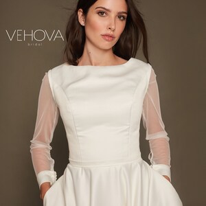 Puede incluir: Un vestido de novia blanco con un corpi&ntilde;o ajustado y una falda completa. El vestido tiene mangas largas de tela transparente. La modelo lleva el vestido y mira a la c&aacute;mara.