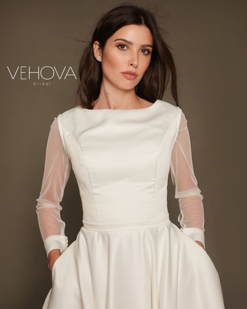 Puede incluir: Un vestido de novia blanco con un corpi&ntilde;o ajustado y una falda completa. El vestido tiene mangas largas de tela transparente. La modelo lleva el vestido y mira a la c&aacute;mara.