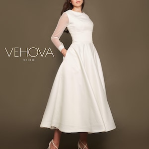 Puede incluir: Un vestido de novia blanco con mangas largas y falda completa. Las mangas son transparentes y el vestido tiene un corpi&ntilde;o ajustado. El vestido est&aacute; fotografiado en una modelo sobre un fondo marr&oacute;n. El texto "VEHOVA bridal" es visible en la imagen.