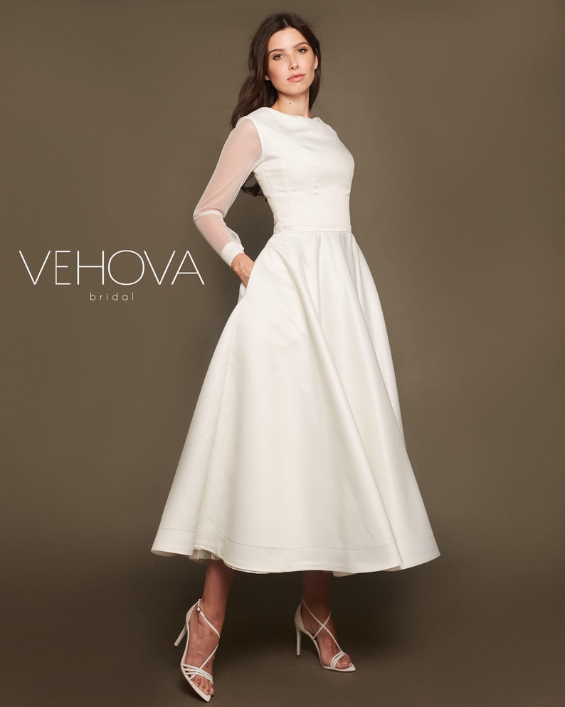 Puede incluir: Un vestido de novia blanco con mangas largas y falda completa. Las mangas son transparentes y el vestido tiene un corpi&ntilde;o ajustado. El vestido est&aacute; fotografiado en una modelo sobre un fondo marr&oacute;n. El texto "VEHOVA bridal" es visible en la imagen.