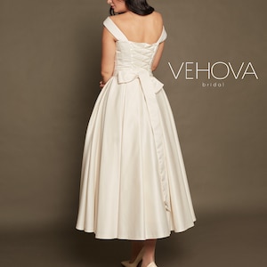 Peut inclure: Une robe de mari&eacute;e en satin blanc avec un d&eacute;collet&eacute; &eacute;paules d&eacute;nud&eacute;es et un dos lac&eacute;. La robe a une jupe ample et un n&oelig;ud dans le dos. La robe est pr&eacute;sent&eacute;e sur un mannequin avec des talons rouges. Le texte "VEHOVA bridal" est visible sur l'image.