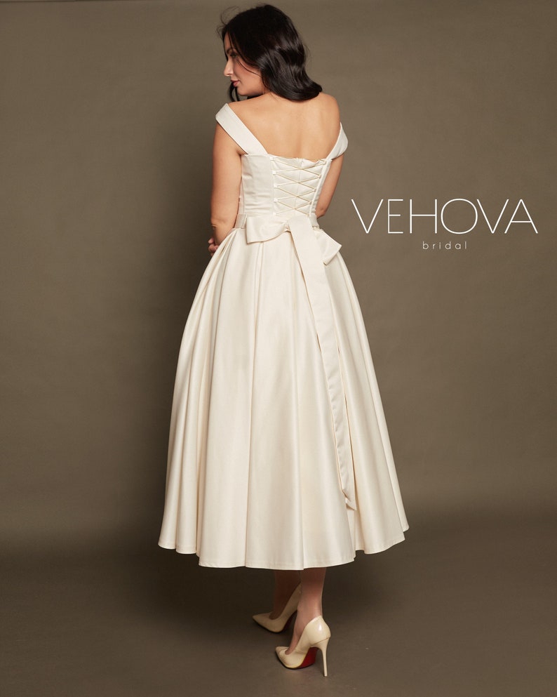 Peut inclure: Une robe de mari&eacute;e en satin blanc avec un d&eacute;collet&eacute; &eacute;paules d&eacute;nud&eacute;es et un dos lac&eacute;. La robe a une jupe ample et un n&oelig;ud dans le dos. La robe est pr&eacute;sent&eacute;e sur un mannequin avec des talons rouges. Le texte "VEHOVA bridal" est visible sur l'image.