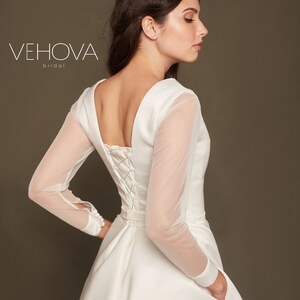 Puede incluir: Un vestido de novia blanco con la espalda de encaje y mangas largas transparentes. El vestido tiene un dise&ntilde;o simple y elegante y una falda fluida.
