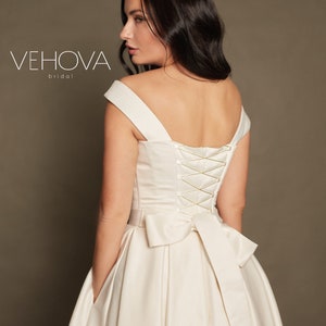 Peut inclure: Une robe de mari&eacute;e en satin blanc avec un d&eacute;collet&eacute; en c&oelig;ur et un dos lac&eacute;. La robe a un grand n&oelig;ud dans le dos et des poches sur les c&ocirc;t&eacute;s. La robe est de Vehova Bridal.