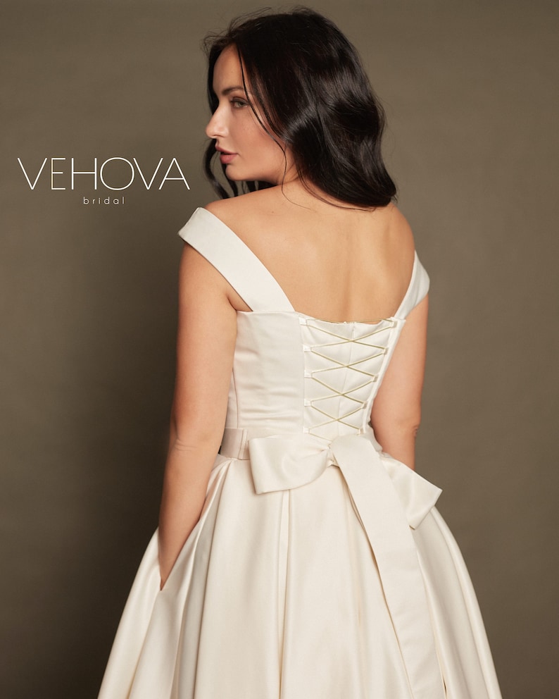 Peut inclure: Une robe de mari&eacute;e en satin blanc avec un d&eacute;collet&eacute; en c&oelig;ur et un dos lac&eacute;. La robe a un grand n&oelig;ud dans le dos et des poches sur les c&ocirc;t&eacute;s. La robe est de Vehova Bridal.