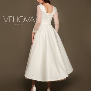 Puede incluir: Un vestido de novia blanco con la espalda con cordones y mangas largas transparentes. La falda es de l&iacute;nea A y llega a la mitad de la pantorrilla. El vestido es de Vehova Bridal.