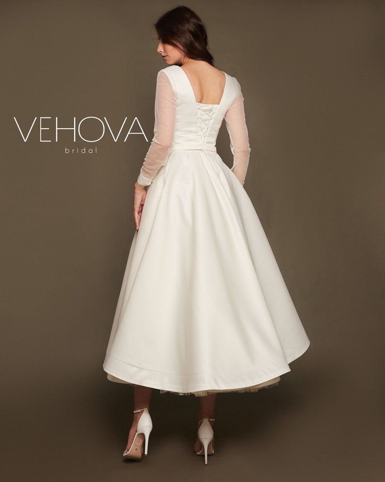 Puede incluir: Un vestido de novia blanco con la espalda con cordones y mangas largas transparentes. La falda es de l&iacute;nea A y llega a la mitad de la pantorrilla. El vestido es de Vehova Bridal.