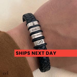 May include: A black braided leather bracelet featuring four silver-toned metal beads. The beads are engraved with an anchor symbol, the name Noah followed by an X, the Roman numeral date V.IX.MMXXI, and an infinity symbol, separated by black rings.