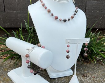 May include: A set of jewelry featuring a necklace, bracelet, and earrings made with pink and brown gemstone beads. The jewelry is displayed on white stands against a brown wooden background.