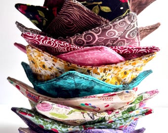 May include: A stack of ten colorful fabric bowl cozies. The cozies are made of different patterned fabrics, including floral, paisley, and geometric designs. The cozies are folded in a triangular shape and stacked on top of each other.