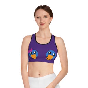 May include: Purple sports bra with a black trim and a cartoon duck design. The duck design features blue heads, orange beaks, and white eyes. One duck has a pink bow. The bra is worn by a person.