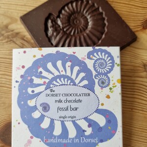 May include: A dark brown chocolate bar shaped like a fossil, alongside a box of chocolate. The box features a blue and white design and the text "The Dorset Chocolatier milk chocolate fossil bar handmade in Dorset".