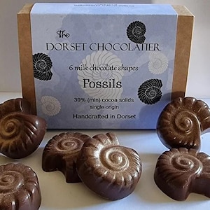 May include: A rectangular cardboard box labelled The Dorset Chocolatier containing 6 milk chocolate shapes in the form of fossils. The packaging features a light blue background with black ammonite illustrations and text stating 39% cocoa solids.