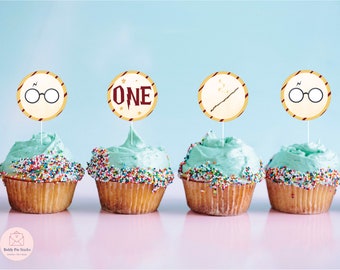 Op de afbeelding: Cupcakes met blauwe glazuur en kleurrijke hagelslag, bekroond met ronde toppers met een bril, een toverstaf en het woord "ONE" in een Harry Potter-thema. De cupcakes staan op een roze ondergrond tegen een blauwe achtergrond.