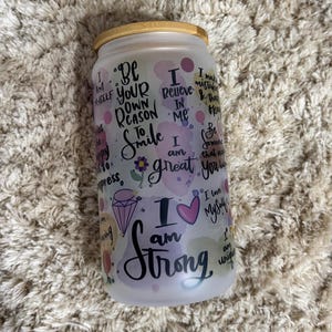 Puede incluir: Un vaso de vidrio esmerilado blanco con tapa de bambú. El vaso tiene un fondo de estilo acuarela con tonos rosados, morados y azules. Presenta varias citas inspiradoras en letras negras, incluyendo "Soy fuerte", "Sé tu propia razón para sonreír" y "Creo en mí".
