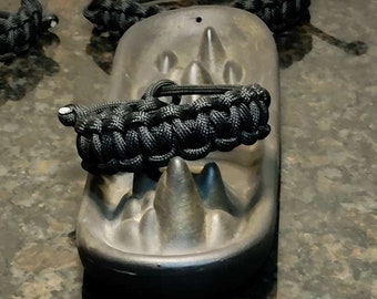 May include: A black paracord bracelet with a textured, dark gray, oval-shaped object. The bracelet is wrapped around the object.