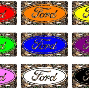 May include: A collection of nine license plates featuring the Ford logo in various colors. Each plate has a camouflage background and the Ford logo in an oval shape. Colors include red, orange, yellow, green, blue, purple, pink, white, and black.