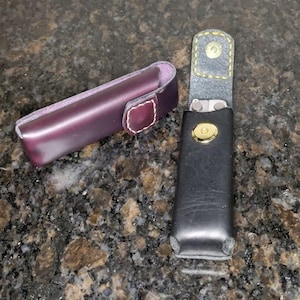 Może przedstawiać: Dwa skórzane etui. Jedno jest w kolorze głębokiego fioletu z białym, szytym akcentem i zapięciem na zatrzask. Drugie jest czarne, otwarte i ma złote zapięcie na zatrzask. Czarne etui ma żółte szwy.