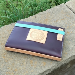 Puede incluir: Un diario encuadernado en cuero marrón con cierre elástico azul. El diario tiene un lomo de cuero marrón claro con la palabra "NOTES" en relieve. La portada presenta un parche de cuero marrón claro con un diseño geométrico.