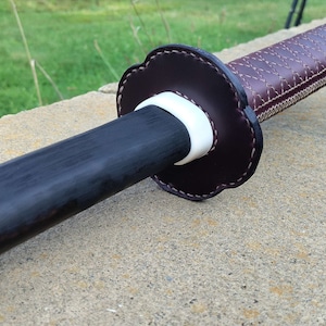 Peut inclure: Gros plan sur une poignée d'épée en bois brun foncé avec un espaceur blanc et un enroulement en cuir brun avec des coutures décoratives.