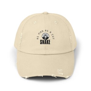 Könnte beinhalten: Beige Baseball Cap mit einem abgenutzten Look und einem gebogenen Schirm. Auf der Kappe befindet sich eine runde Grafik mit einer Schlange und dem Text "AS MAD AS A CUT SNAKE" in Schwarz. Vintage-Look.