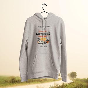 May include: A light gray hoodie with a front pocket and a drawstring hood. The front graphic reads "John & Julie are Living the Grey Nomad Dream" with a camper van illustration and "Est. 2026". The hoodie is hanging on a black hanger.