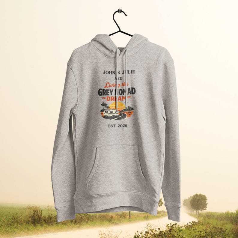 May include: A light gray hoodie with a front pocket and a drawstring hood. The front graphic reads "John & Julie are Living the Grey Nomad Dream" with a camper van illustration and "Est. 2026". The hoodie is hanging on a black hanger.
