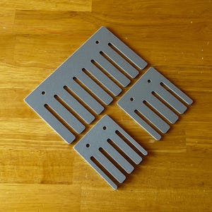 Può includere: Quattro oggetti grigi a forma di pettine con denti scanalati e piccoli fori, disposti su una superficie di legno. Gli oggetti sono di dimensioni variabili, con uno più grande in alto e tre più piccoli sotto.