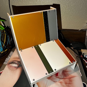 May include: A multi-colored, rectangular, hinged box with a white frame. The box features panels in gold, black, gray, pink, olive green, white, and orange. The box is held in a person's hand.