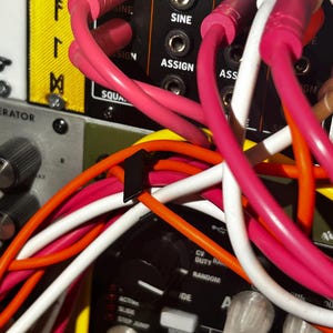 Peut inclure: Gros plan sur un panneau de synth&eacute;tiseur modulaire avec divers c&acirc;bles de raccordement color&eacute;s branch&eacute;s. Les c&acirc;bles sont roses, blancs, oranges et jaunes. Le panneau porte les mentions "RESET/SYNC", "SINE" et "ASSIGN".