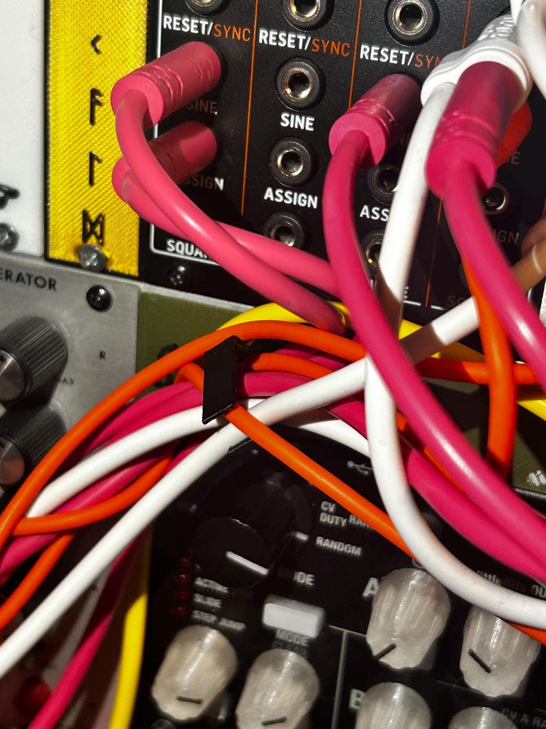 Peut inclure: Gros plan sur un panneau de synth&eacute;tiseur modulaire avec divers c&acirc;bles de raccordement color&eacute;s branch&eacute;s. Les c&acirc;bles sont roses, blancs, oranges et jaunes. Le panneau porte les mentions "RESET/SYNC", "SINE" et "ASSIGN".