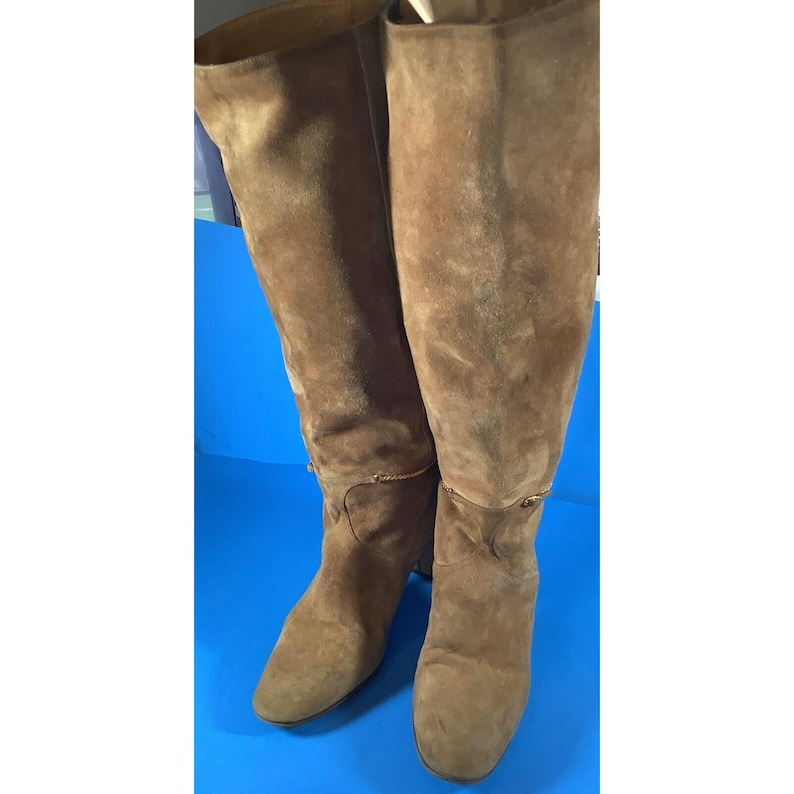 May include: A pair of knee-high brown suede boots. The boots have a pointed toe and a small heel. A gold-colored chain detail is visible around the top of the boot. The boots are displayed against a blue background.