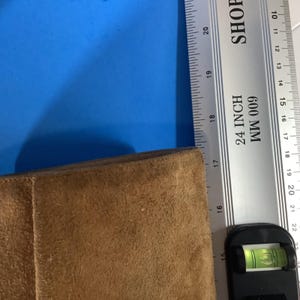 May include: A tan suede material is measured with a silver ruler marked in inches and millimeters. The ruler reads "24 INCH 600 MM". A level is at the bottom of the image. The background is blue.