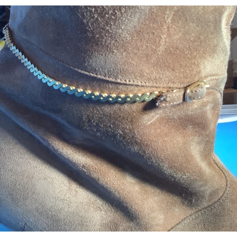 May include: A brown suede boot with a gold-colored chain ankle bracelet. The ankle bracelet is a delicate chain with a repeating pattern. The boot has a buckle detail and a soft, textured appearance.