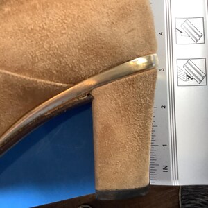 May include: Close-up of a tan suede heeled shoe with a gold-colored trim. The heel is approximately 2 inches tall, as measured by the ruler in the image. The shoe is set against a blue and brown background.