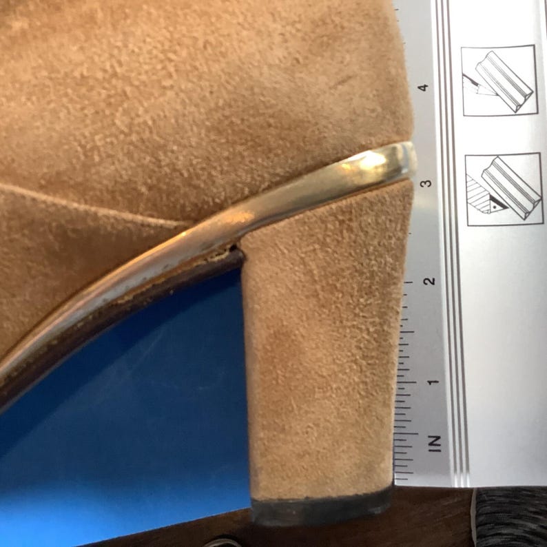 May include: Close-up of a tan suede heeled shoe with a gold-colored trim. The heel is approximately 2 inches tall, as measured by the ruler in the image. The shoe is set against a blue and brown background.
