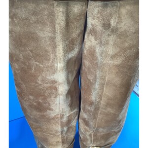 May include: Close-up of two brown suede boots. The boots have a knee-high design and a textured surface. The boots have a gold chain detail at the bottom. The background is a bright blue.