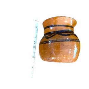 Könnte beinhalten: Kleiner, brauner Keramiktopf mit schwarz bemalten Bändern und dekorativen Schnörkeln. Der Topf ist etwa 10 cm hoch und hat einen abgerundeten Körper mit einem leicht ausgestellten Rand. Der Topf ist glasiert und hat eine glänzende Oberfläche.