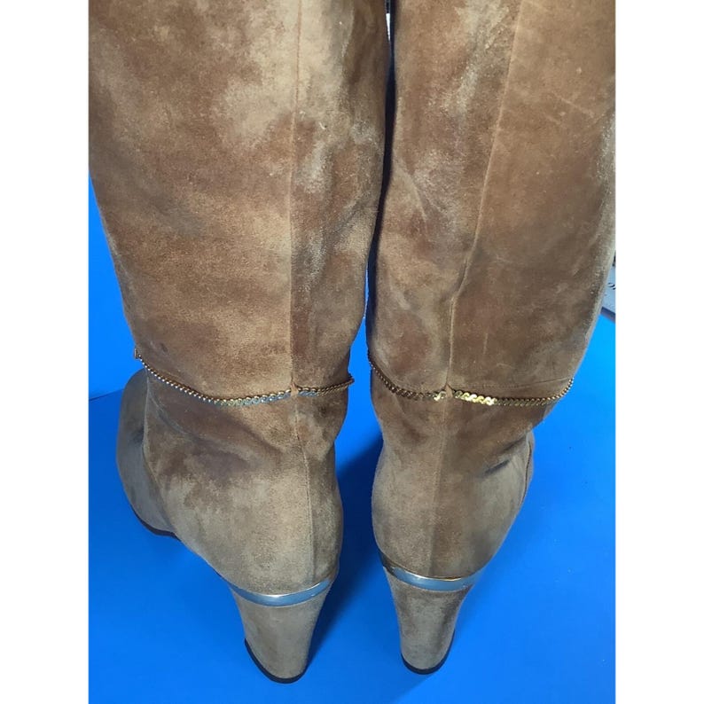 May include: A pair of knee-high brown suede boots. The boots have a gold chain detail around the top and silver trim on the heels. The heels are approximately 3 inches high.