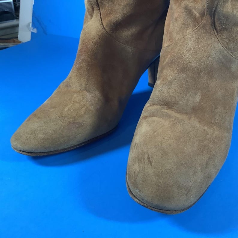 May include: A pair of brown suede boots with a pointed toe and a mid-height heel. The boots have a soft, textured appearance and are set against a blue background. The boots appear to be in good condition.