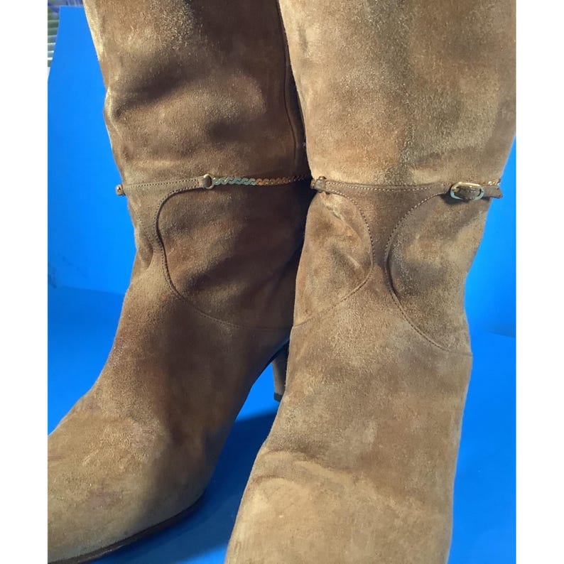 May include: A pair of knee-high brown suede boots. The boots have a small heel and a decorative strap around the calf. The suede has a soft, textured appearance. The boots are set against a blue background.