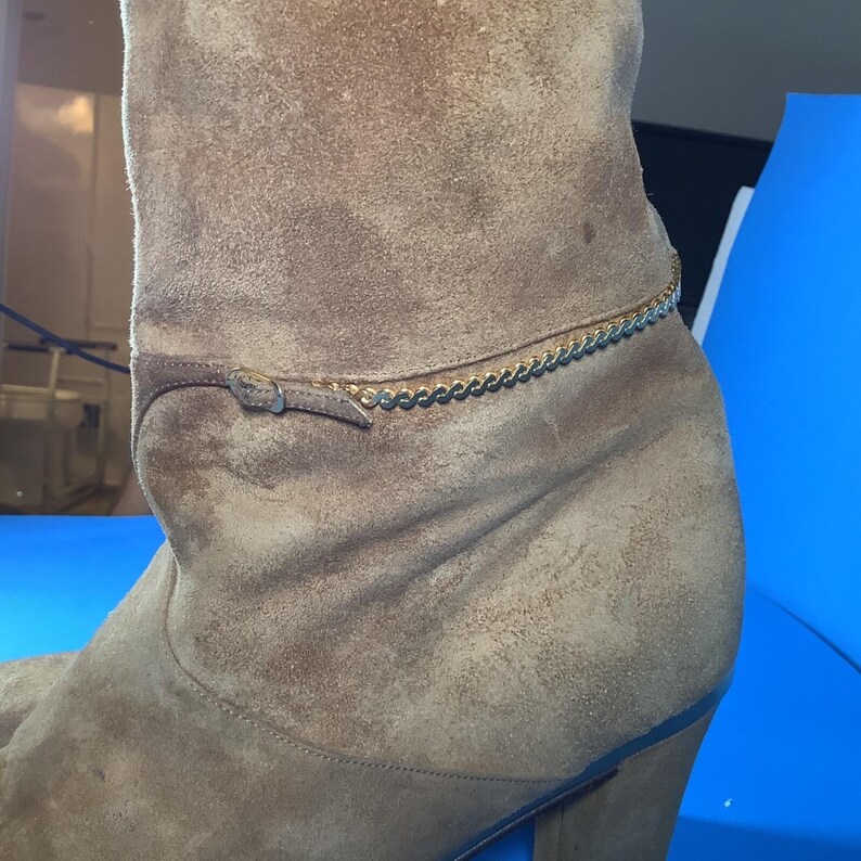 May include: A tan suede boot with a gold chain ankle detail. The boot has a brown leather strap with a small buckle. The heel is a dark brown color.