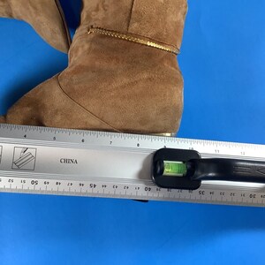 May include: A pair of brown suede boots with a gold trim. The boots are measured with a ruler, showing a length of approximately 10 inches. The boots are on a blue surface.