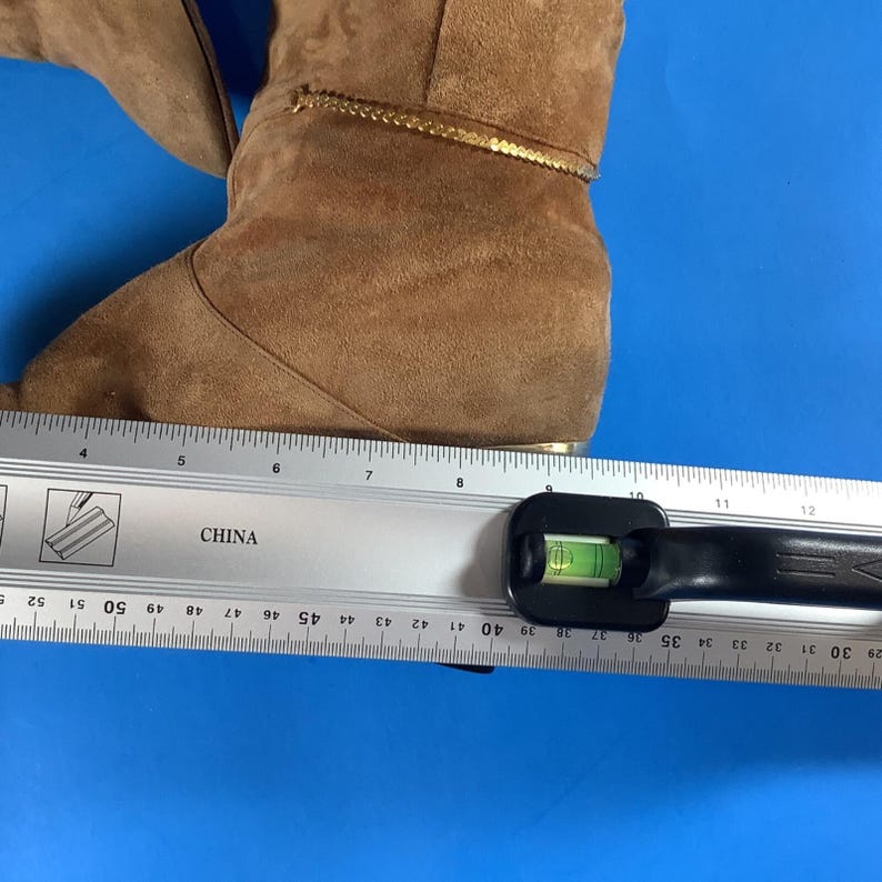 May include: A pair of brown suede boots with a gold trim. The boots are measured with a ruler, showing a length of approximately 10 inches. The boots are on a blue surface.