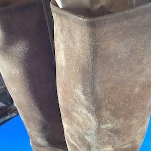 May include: Close-up of two brown suede boots. The boots have a tall shaft and a soft, textured appearance. The color is a warm, earthy brown, and the suede has a slightly worn look. The boots appear to be unworn and in good condition.