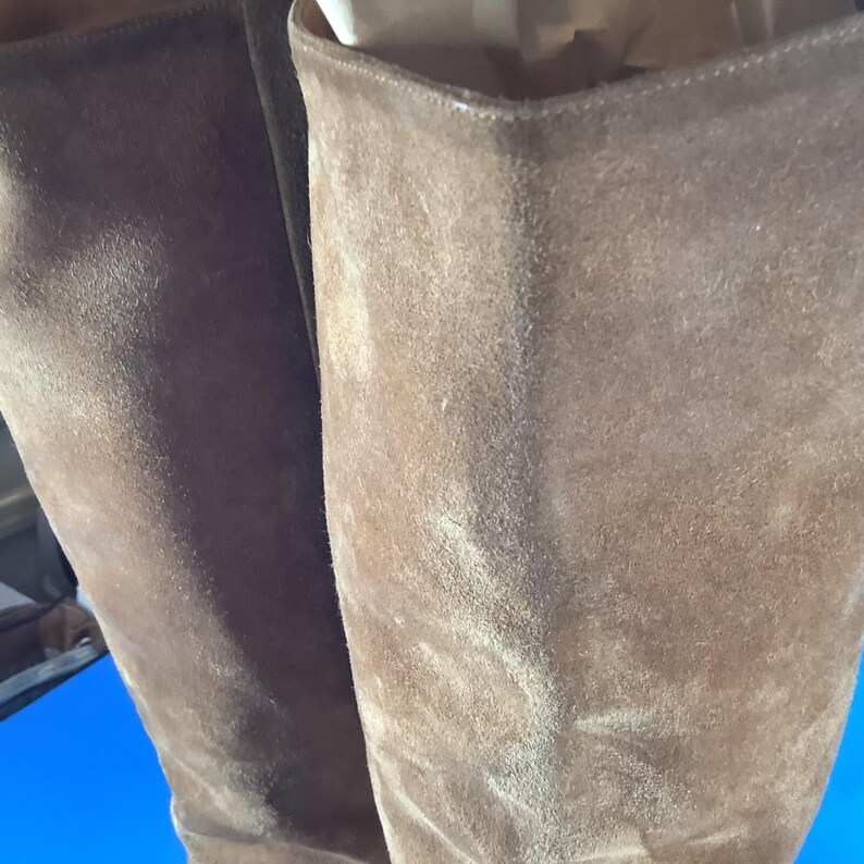 May include: Close-up of two brown suede boots. The boots have a tall shaft and a soft, textured appearance. The color is a warm, earthy brown, and the suede has a slightly worn look. The boots appear to be unworn and in good condition.