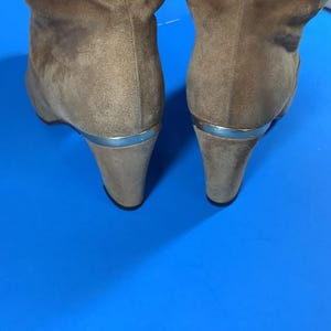 May include: A pair of taupe suede boots with wedge heels. The boots have a silver-colored metal accent at the heel. The background is a bright blue.