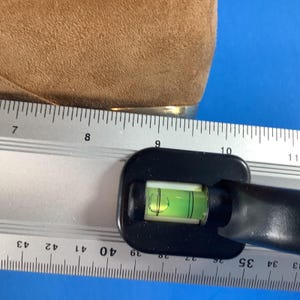 May include: A black level tool with a green bubble is positioned next to a ruler. The ruler shows measurements in inches. A tan suede material is visible in the upper left corner.