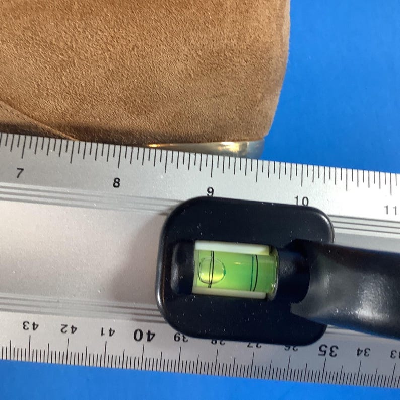 May include: A black level tool with a green bubble is positioned next to a ruler. The ruler shows measurements in inches. A tan suede material is visible in the upper left corner.