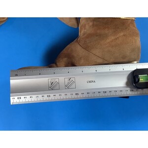 May include: A brown suede boot with a gold-colored trim. The boot is measured with a ruler, showing a length of approximately 7 inches. The word "CHINA" is printed on the ruler.
